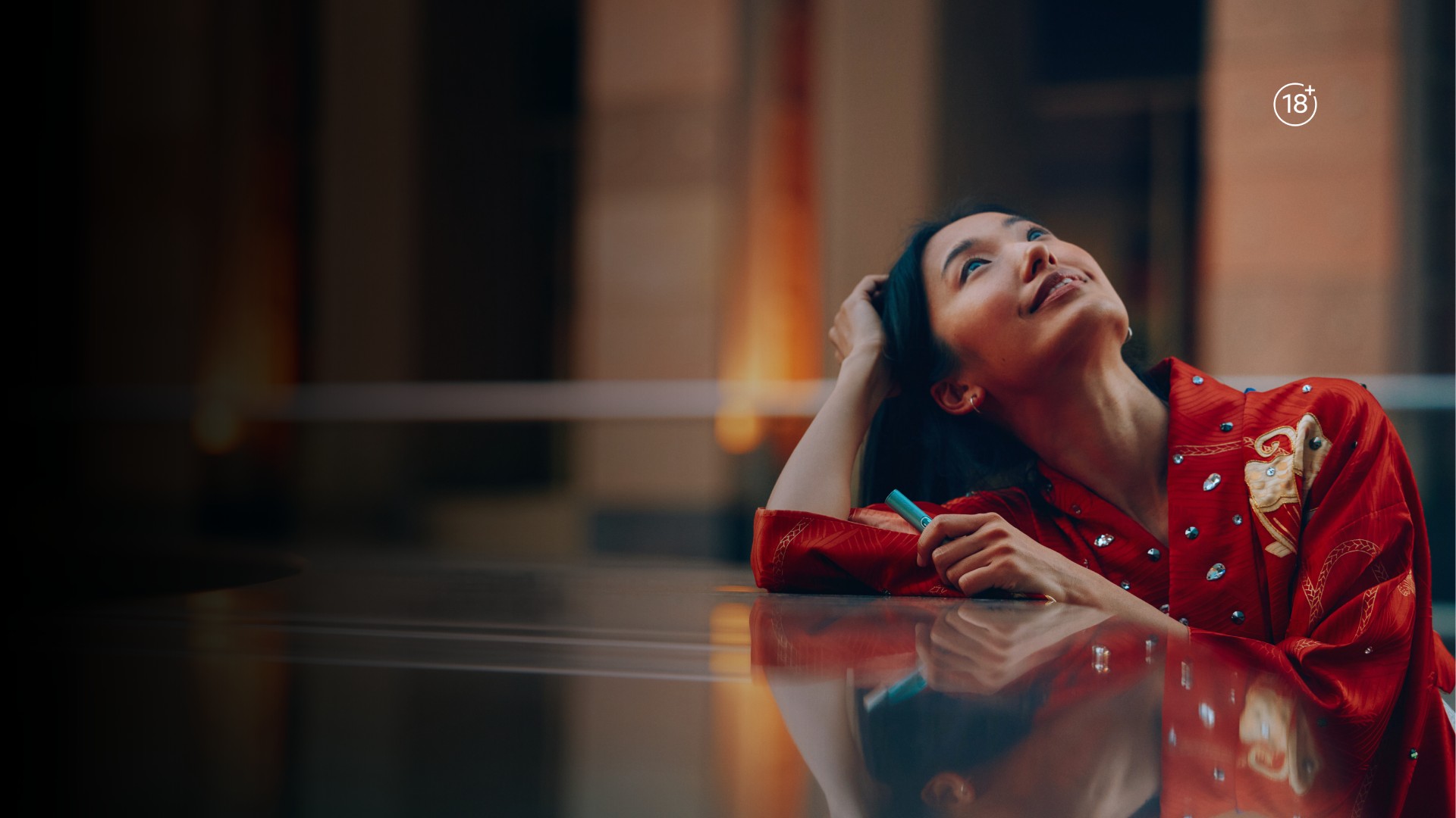 Woman sitting outdoors and looking upward with warm ambient lighting reflecting on a glossy surface.