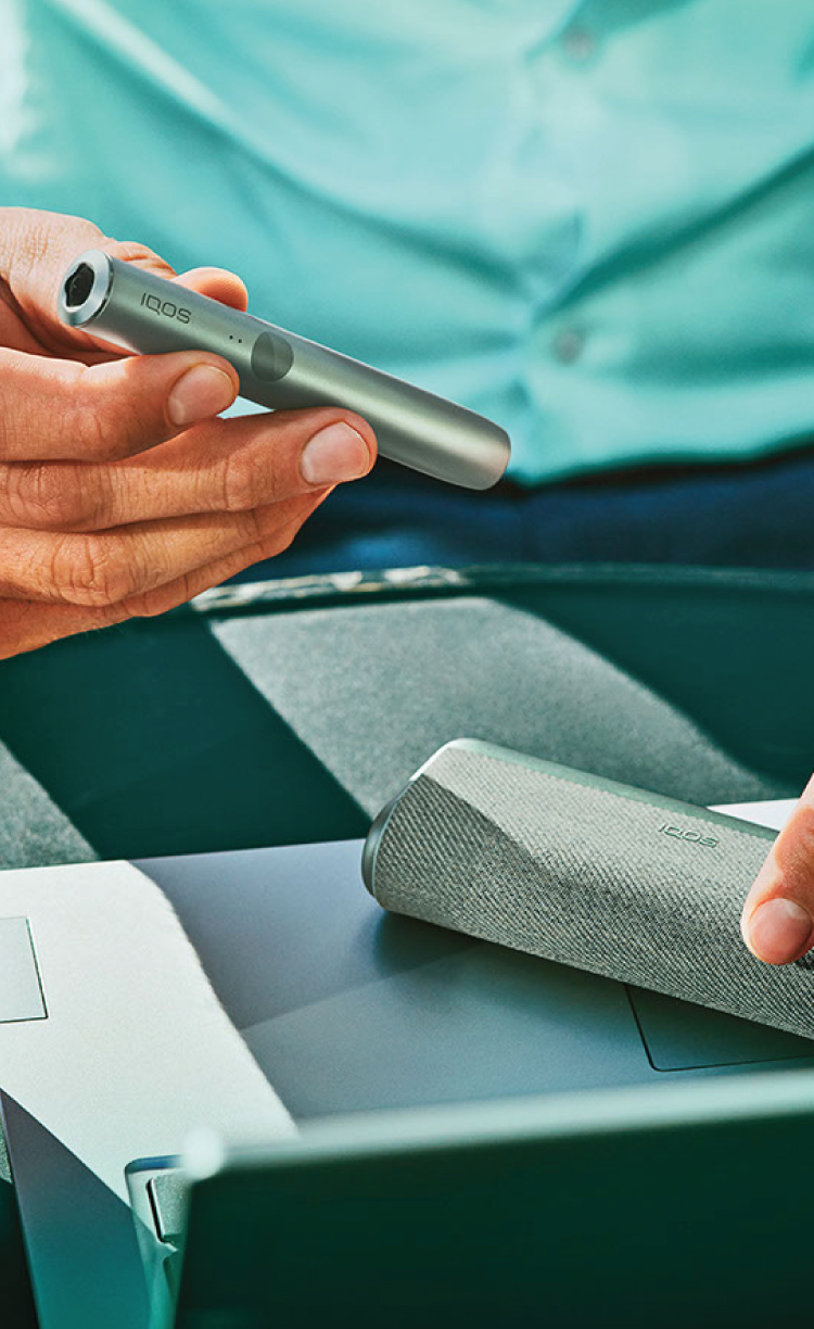 An IQOS device held by one person and a smartphone held by another person.