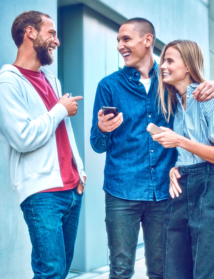 Two people holding IQOS ILUMA devices smile as their friend looks at the IQOS app on her mobile phone.