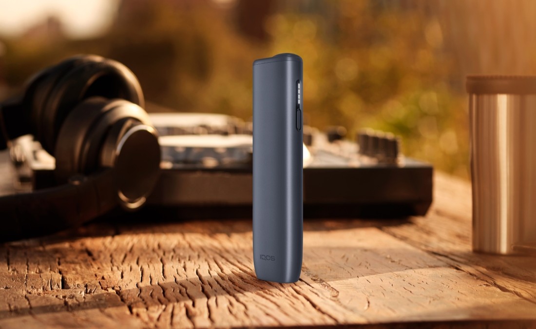 Устройство IQOS ILUMA i ONE цвета Черный графит на деревянном столе с наушниками