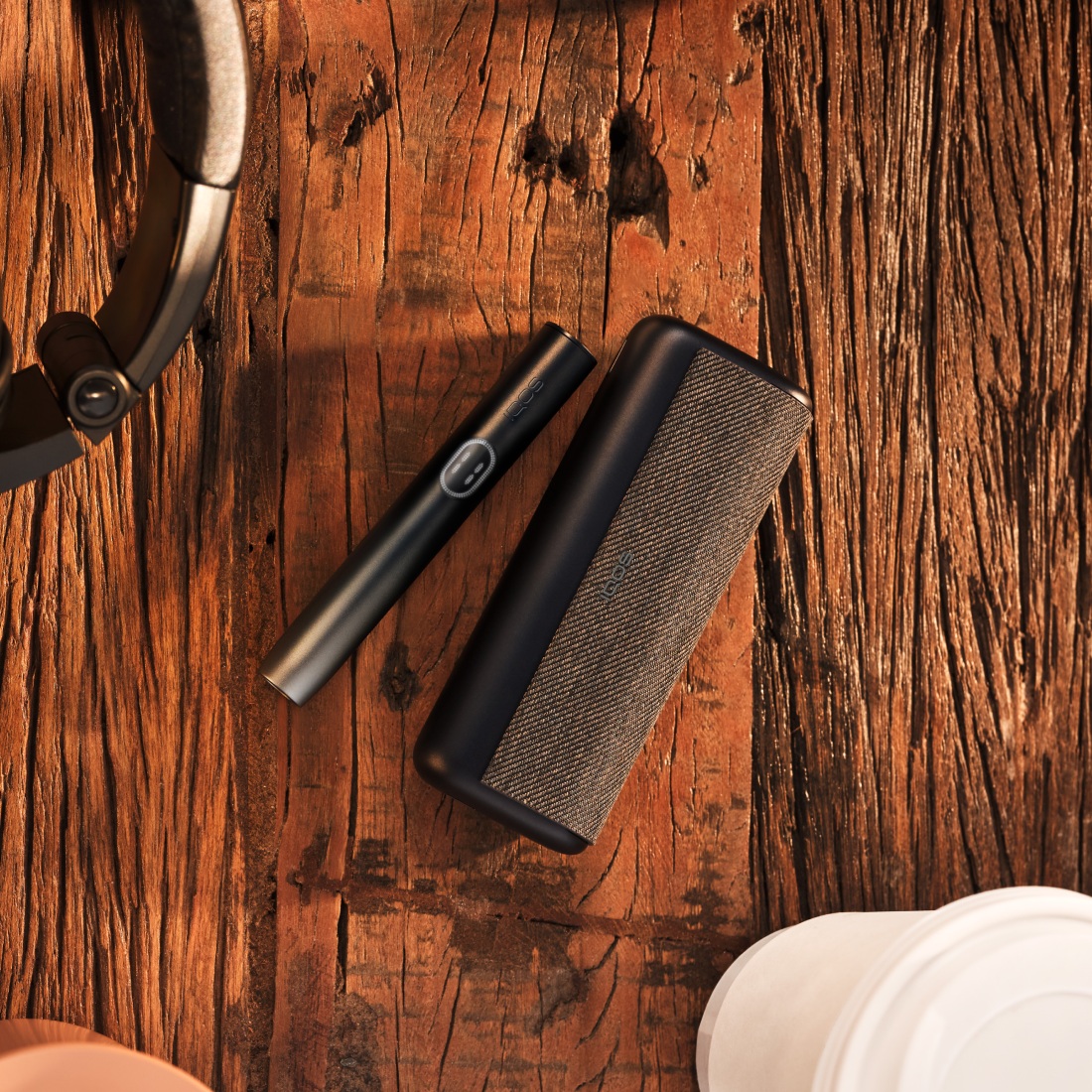 Устройство IQOS ILUMA i PRIME цвета Черный графит на столе с кофе и наушниками