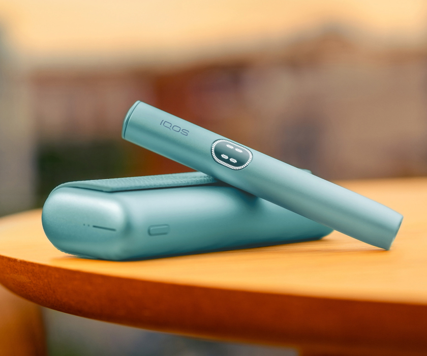 Устройство IQOS ILUMA i PRIME цвета Лазурный бриз над зарядным устройством на столе