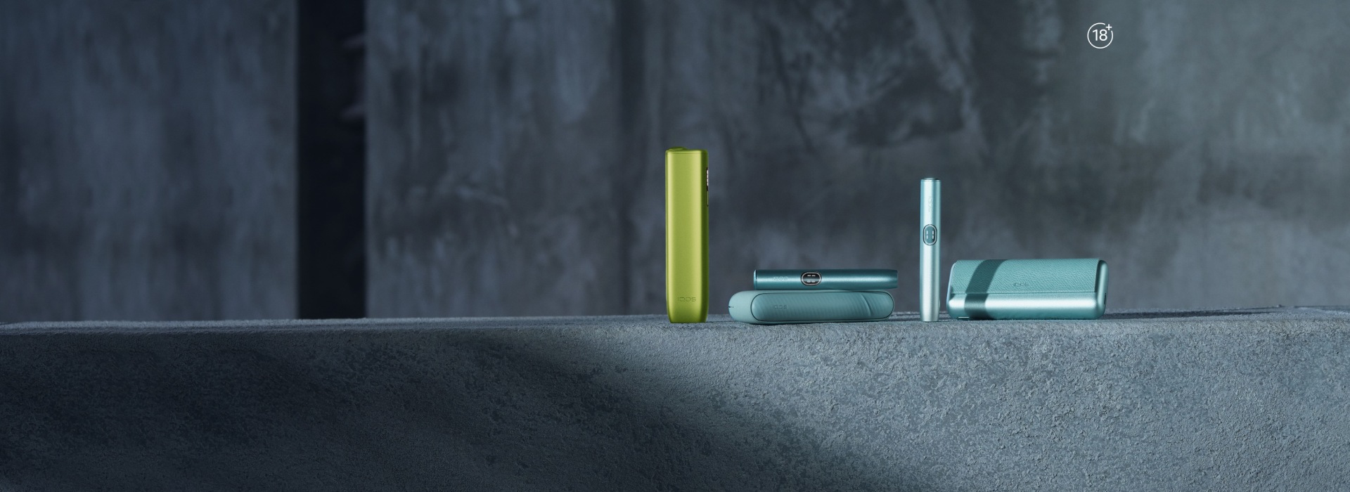 IQOS ILUMA i devices in green and blue displayed on a textured gray surface with a dark concrete background.