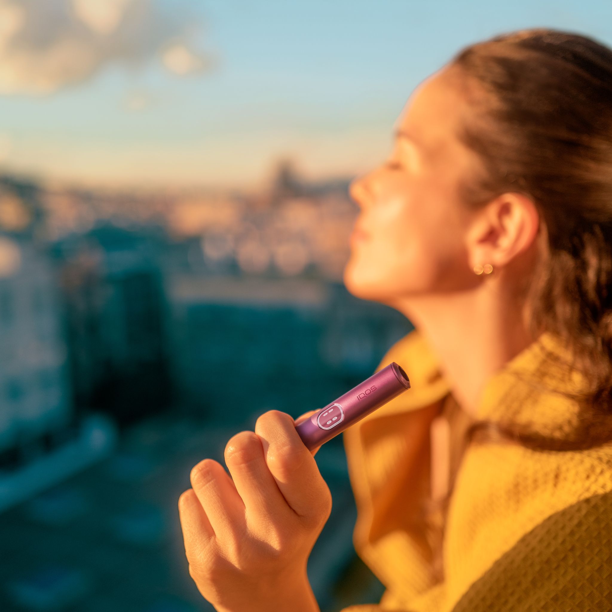 Person in yellow holding a Garned Red IQOS ILUMA i Prime Holder outdoors with city buildings and cloudy sky in the background