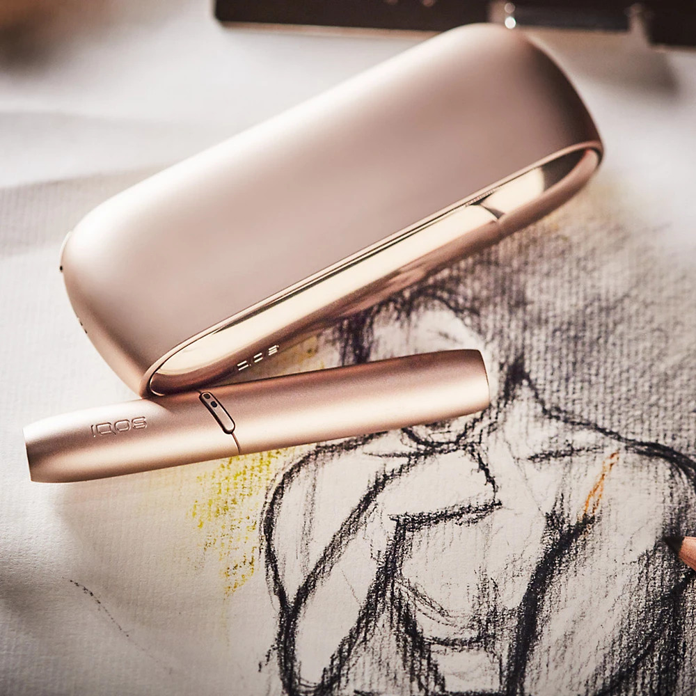 Close-up of a sleek, gold IQOS heated tobacco device and its matching charging case placed on a textured surface with artistic sketches in the background.