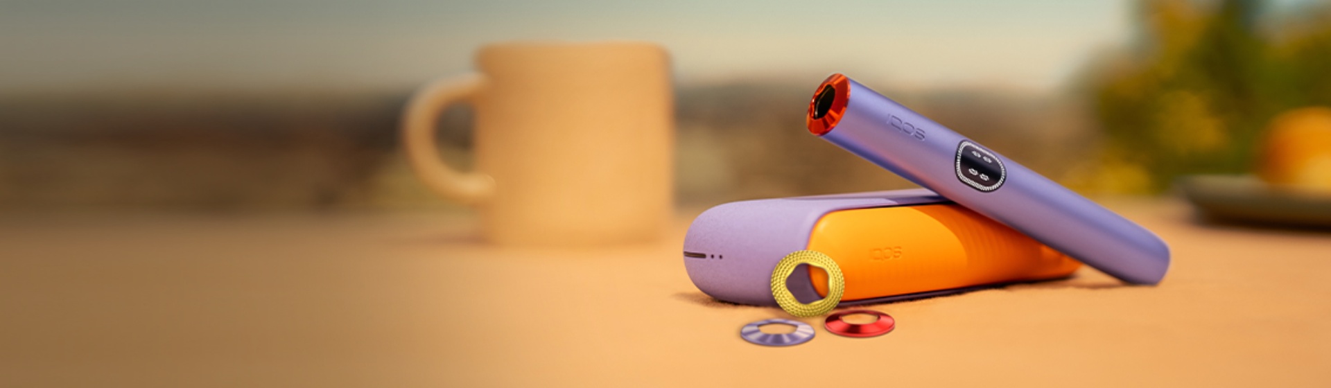 Close-up of Digital Violet IQOS ILUMA i adorned with accessories, placed on a table with a mug and set against a blurred outdoor background.