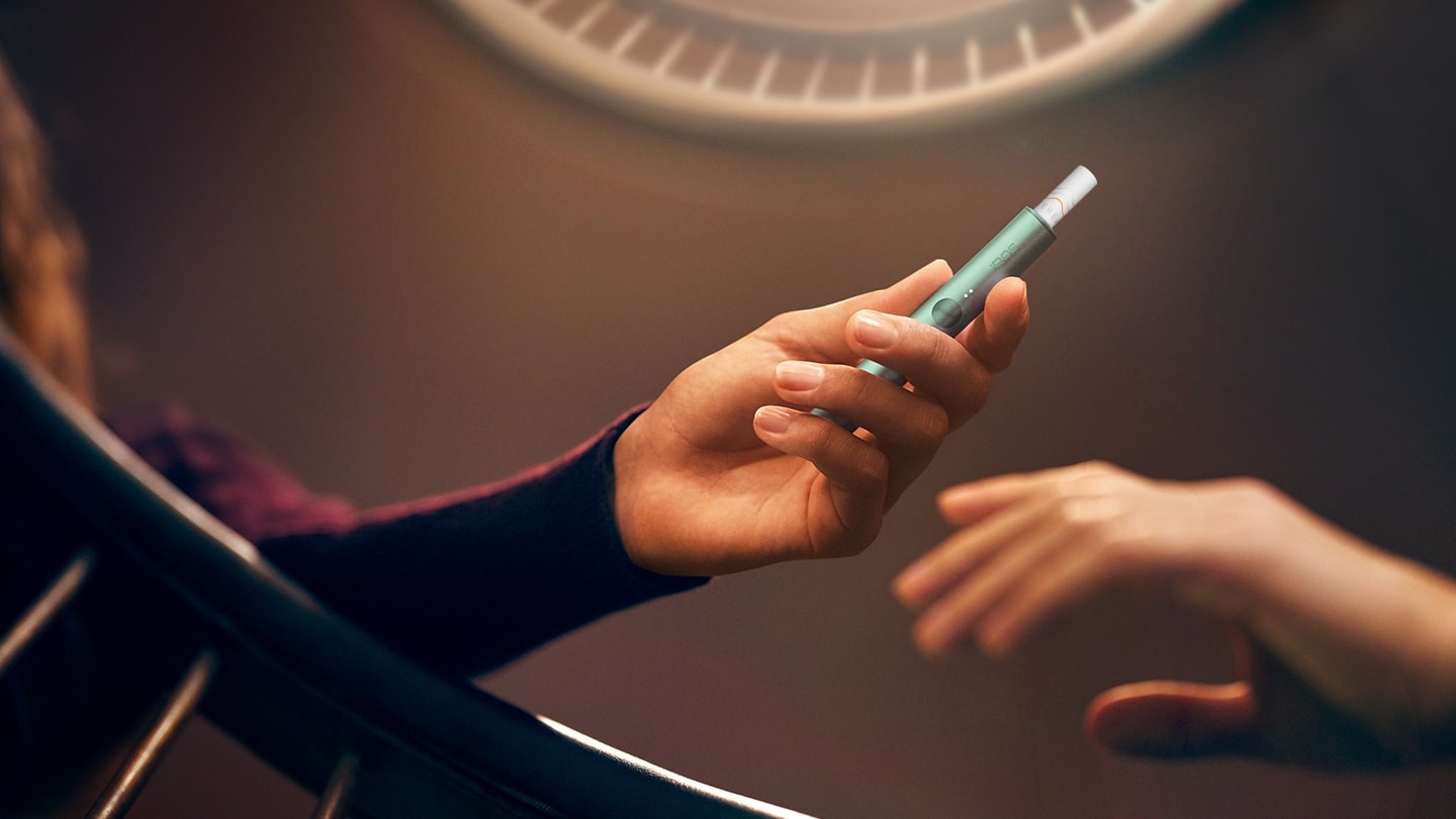 Two hands on a staircase handle. One holding an IQOS ILUMA holder with a TEREA stick.