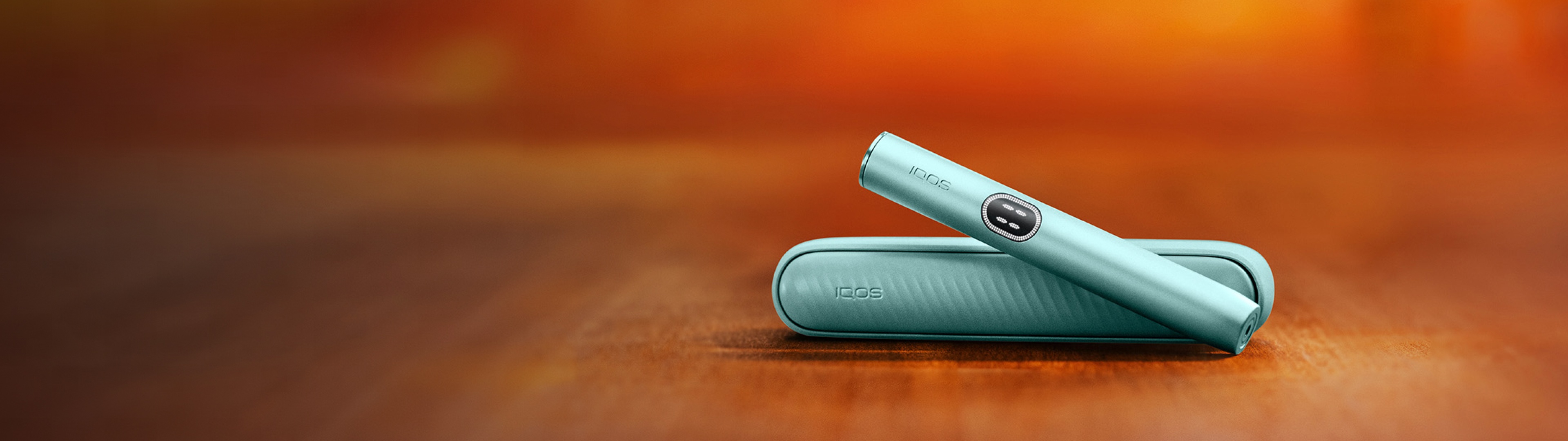 in IQOS ILUMA i Gerät in der Farbe Blue Breeze. Der Hintergrund ist in warmen Orangetönen unscharf gehalten.
