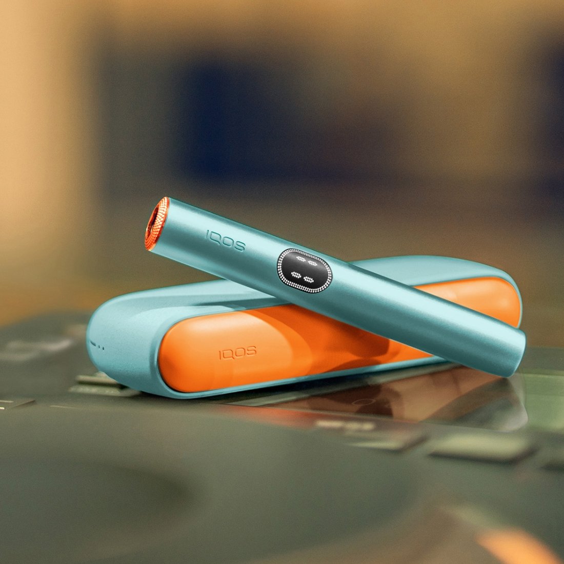 Ein IQOS ILUMA i Gerät in der Farbe Breeze Blue liegt auf einer Oberfläche. Es ist mit einem orangefarbenen Klappen-Cover versehen, und der Holder trägt einen orangefarbenen Ring als Zubehör.