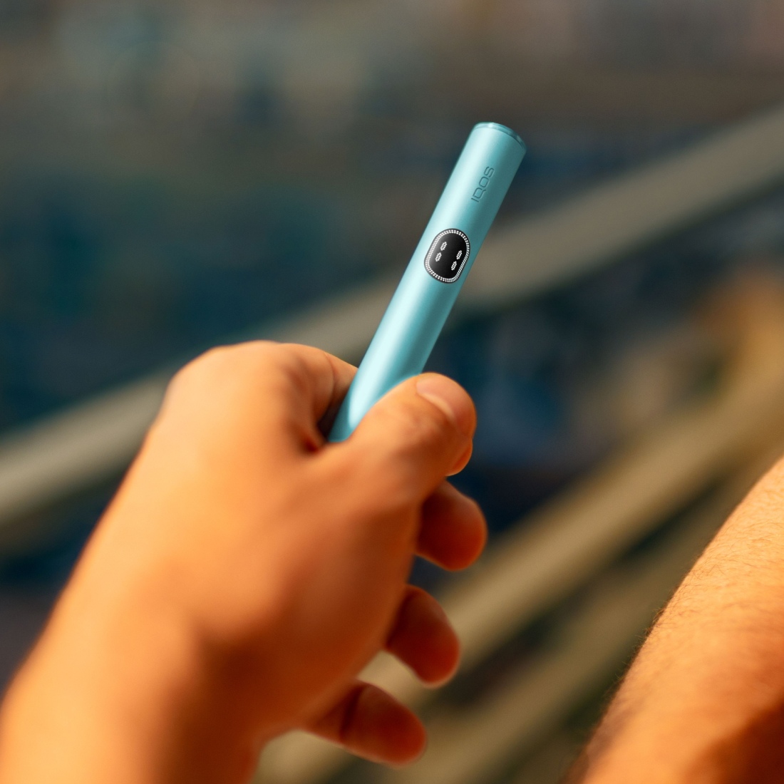 Eine Hand hält den Holder eines IQOS ILUMA i Geräts in der Farbe Breeze Blue. Das Gerät ist zylindrisch, hat eine glatte Oberfläche und trägt den Aufdruck ‚IQOS‘.