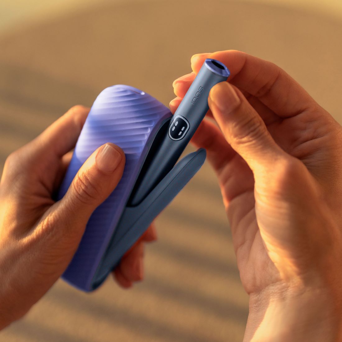 Eine Person hält ein IQOS ILUMA i Gerät in der Farbe Rot. Der Pocket Charger ist mit einer lila Silikonhülle versehen, und der Holder des Geräts trägt einen lila Ring als Zubehör. Die Hände der Person sind sichtbar, während sie mit dem Gerät interagiert.