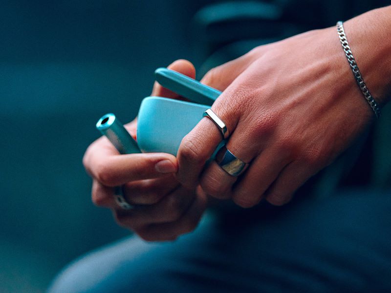 Eine Hand hält ein IQOS ILUMA i Gerät in der Farbe Breeze Blue. Das Gerät ist zylindrisch geformt und hat eine glatte, metallische Oberfläche.