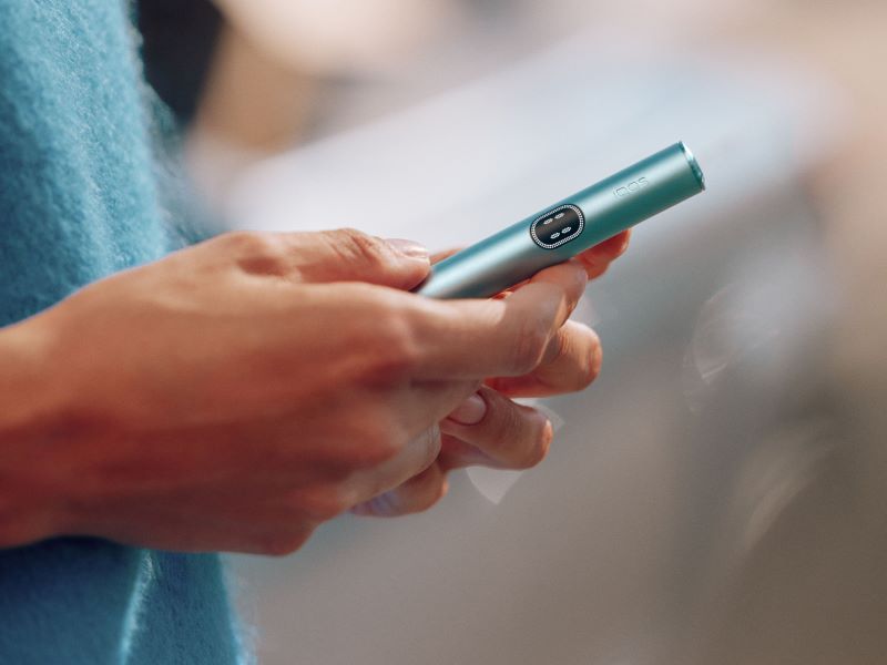 Hände halten einen IQOS ILUMA i Holder in Breeze Blue vor unscharfem Hintergrund.