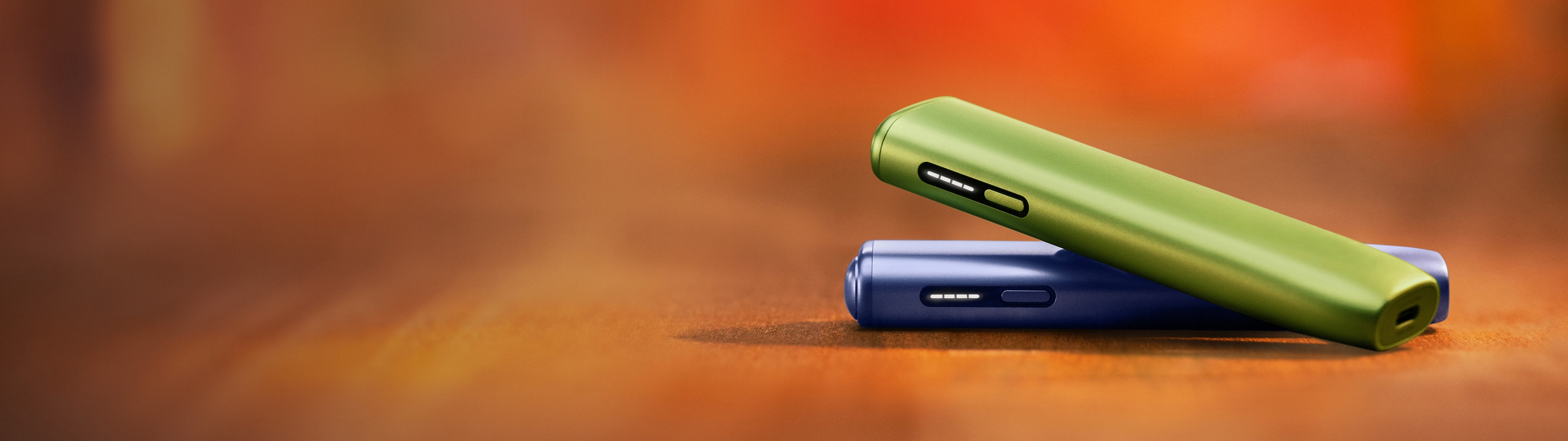 Zwei IQOS ILUMA i ONE Geräte in den Farben Green und Violet liegen übereinander auf einer glatten Oberfläche.