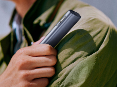 Eine Hand hält ein IQOS ILUMA i ONE Gerät in der Farbe Midnight Black mit abgerundeten Kanten, seitlichem Knopf und LED-Anzeige, vor unscharfem Hintergrund.