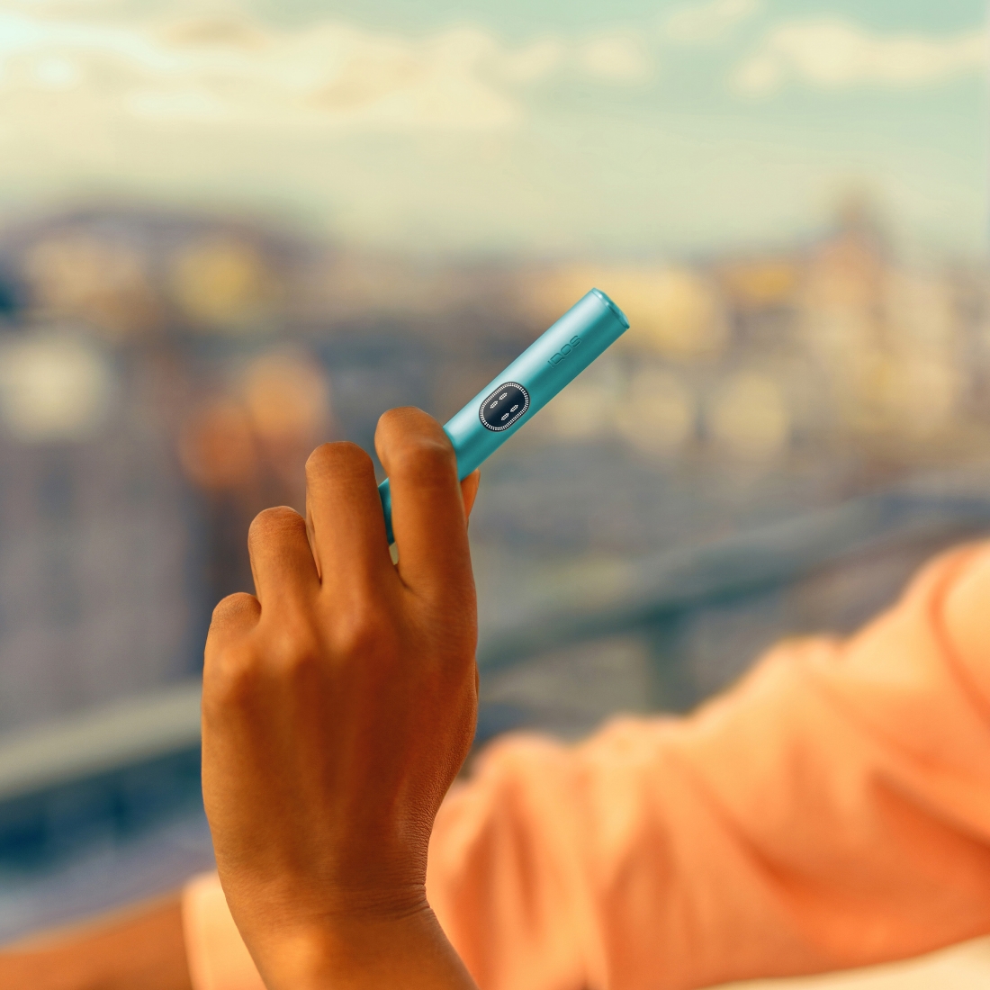 Eine Hand hält den Holder des IQOS ILUMA i Geräts in der Farbe Breeze Blue, vor unscharfem Hintergrund mit Gebäuden und bewölktem Himmel.