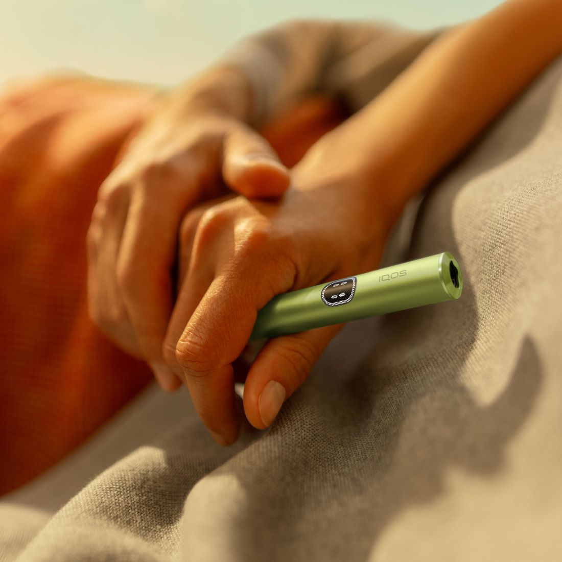 Eine Hand hält den Holder des IQOS ILUMA i i Geräts in der Farbe Green, vor unscharfem Hintergrund.