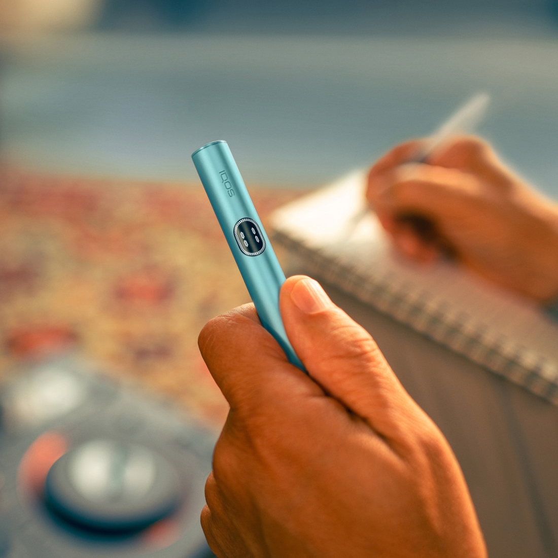 Eine Person hält den Holder des IQOS ILUMA i Geräts in der Farbe Breeze Blue und schreibt gleichzeitig mit einem Stift in ein Notizbuch. Der Hintergrund ist unscharf und zeigt eine Innenraumszene.