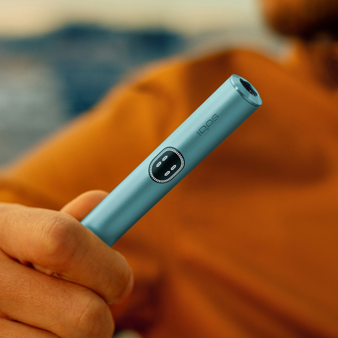 Eine Hand hält den Holder des IQOS ILUMA i Geräts in der Farbe Breeze Blue mit schlankem, zylindrischem Design und kleinem Knopf oder Indikator. Der Hintergrund ist unscharf, die Person trägt ein orange- oder braunfarbenes Kleidungsstück.