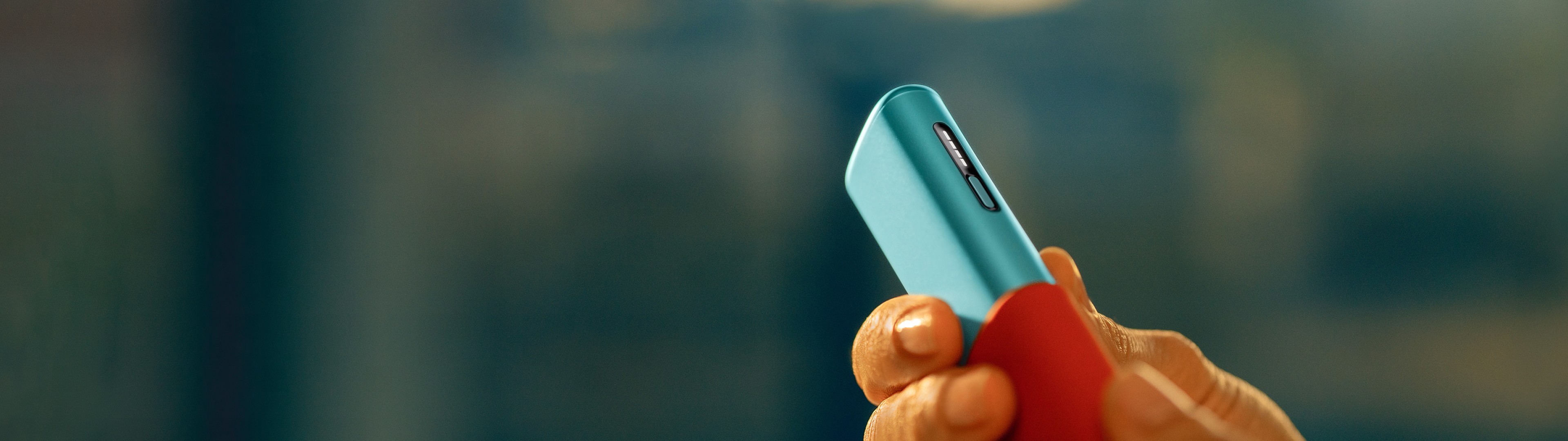 Eine Hand zieht ein blaues IQOS ILUMA i ONE Gerät aus einer roten Schutzhülle.