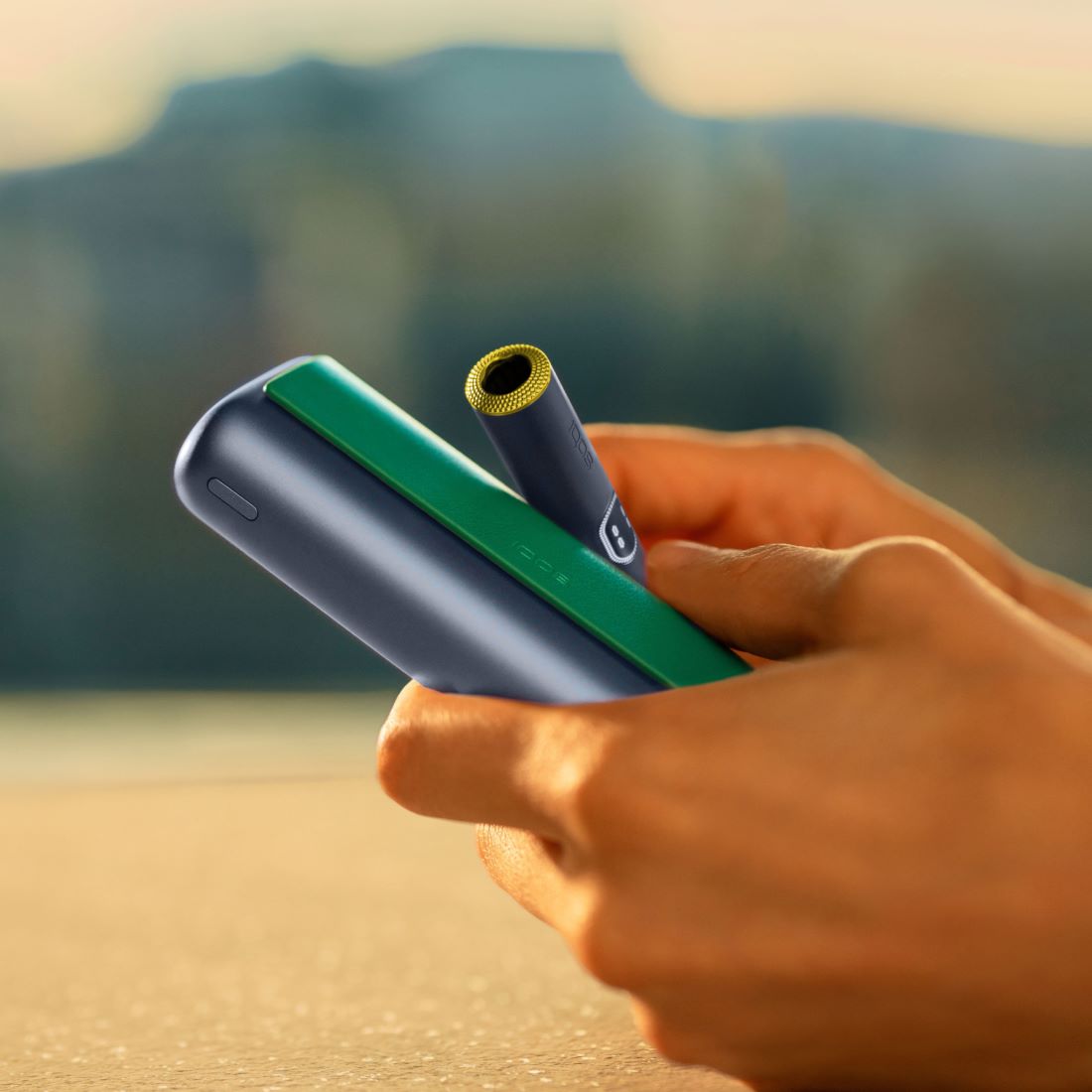 Eine Person hält ein IQOS ILUMA i PRIME Gerät in der Farbe Midnight Black mit grünem Klappen Cover und gelbem Ringzubehör am Holder.