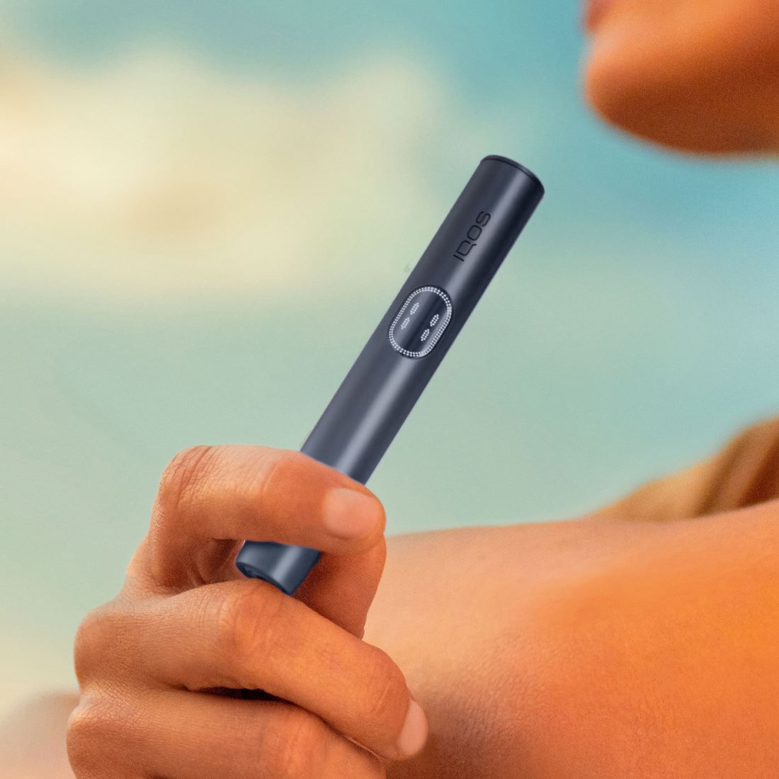 Eine Hand hält den Holder des IQOS ILUMA i PRIME Geräts in der Farbe Midnight in heller Outdoor-Umgebung.