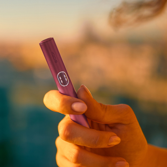 Əlində Garnet red IQOS ILUMA i PRIME cihazı tutan şəxs