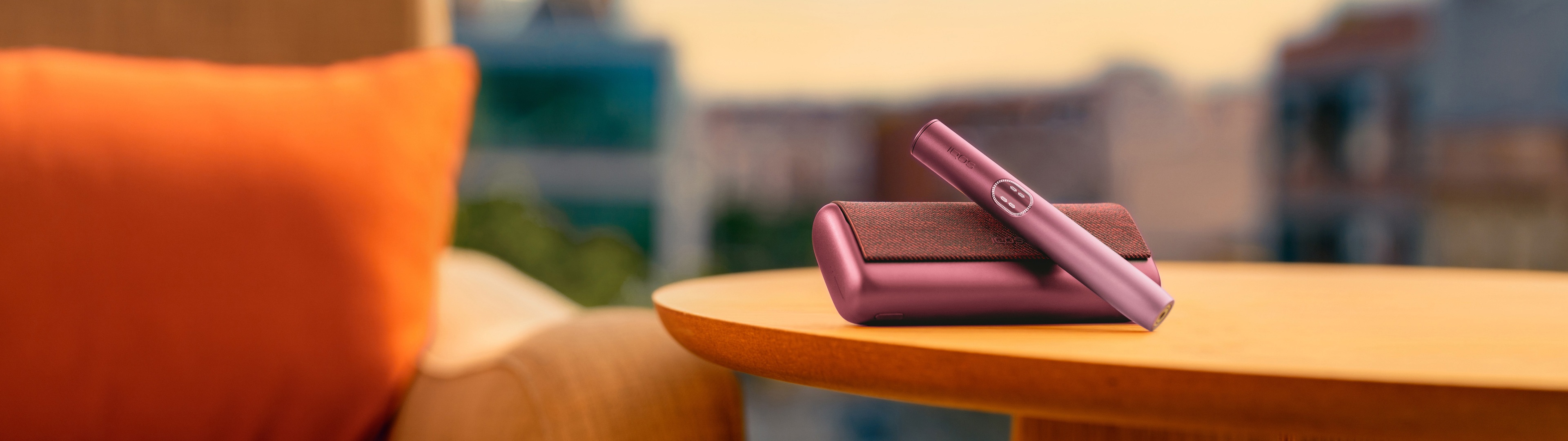 Устройство IQOS ILUMA i PRIME гранатово-красного цвета над зарядным устройством, на столе возле дивана