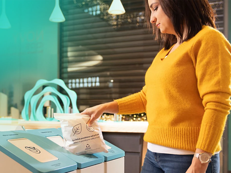 Žena v oranžovém svetru vhazující do recyklačního boxu plný sáček IQOS