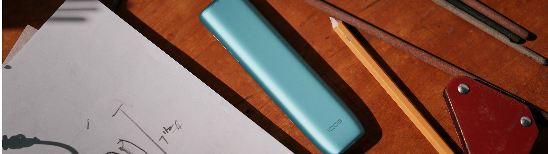 IQOS ILUMA i ONE color Breeze Blue al lado de un cuaderno de dibujo y un lápiz.