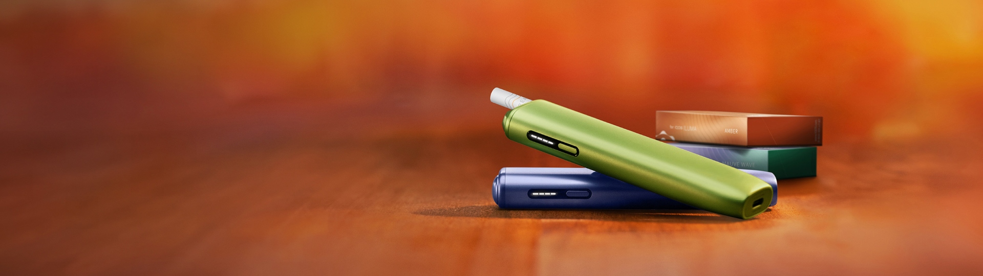 جهاز IQOS ILUMA i ONE أخضر فوق جهاز IQOS ILUMA i ONE لافندر، بالقرب من عبوتين من المواد الاستهلاكية.