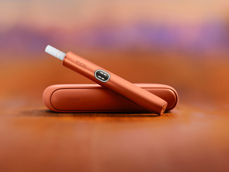 جهاز IQOS ILUMA i باللون الأحمر الطوبي فوق شاحنه، مع خلفية حمراء