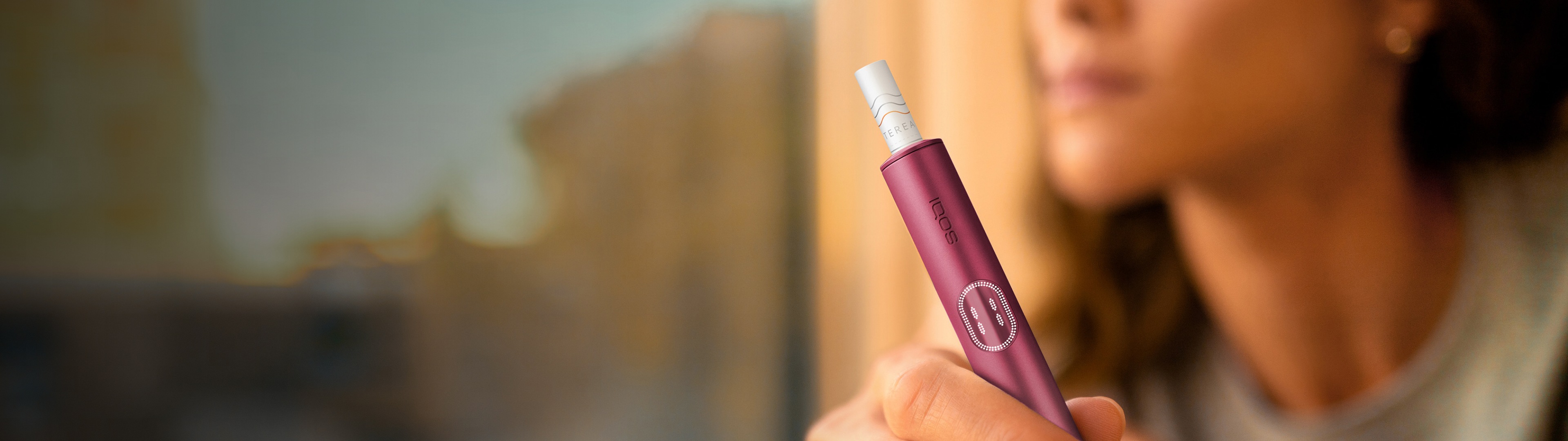 Woman holding a Garnet Red IQOS ILUMA i PRIME device, while looking outside of the window