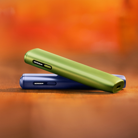 2 appareils IQOS ILUMA i ONE (appareil tout-en-un) couleurs Vert Anis et Lila, posés l'un sur l'autre, sur un fond dégradé orange
