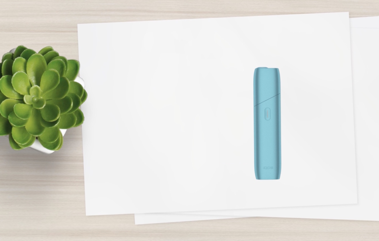 Türkiser IQOS ORIGINALS ONE liegt auf einem weißen Blatt neben einer Pflanze
