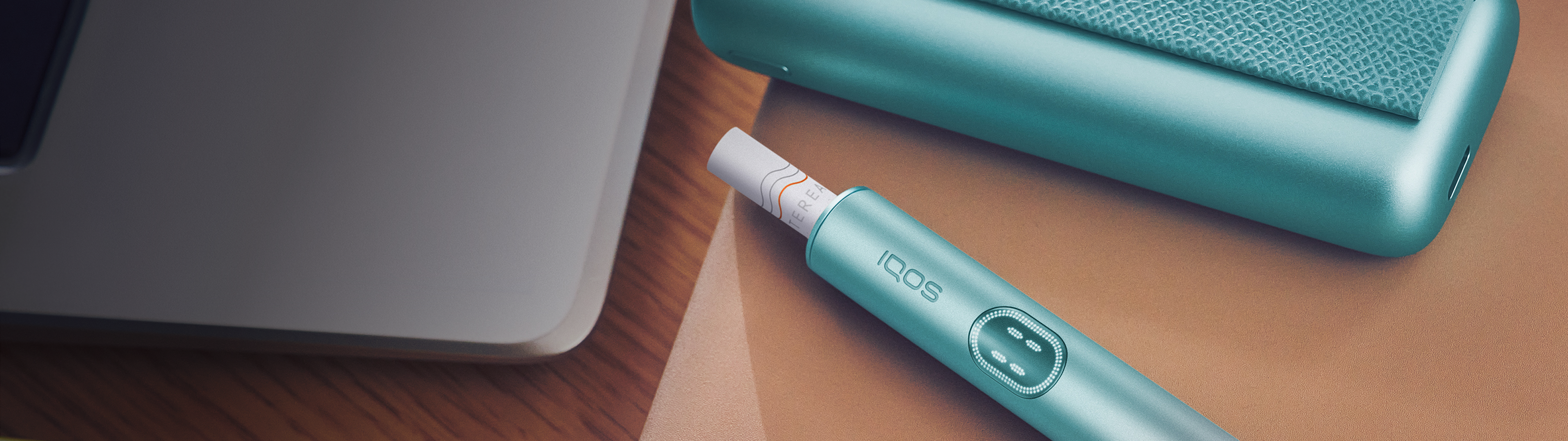 IQOS ILUMA i liegt auf einem Tisch neben einem Laptop.