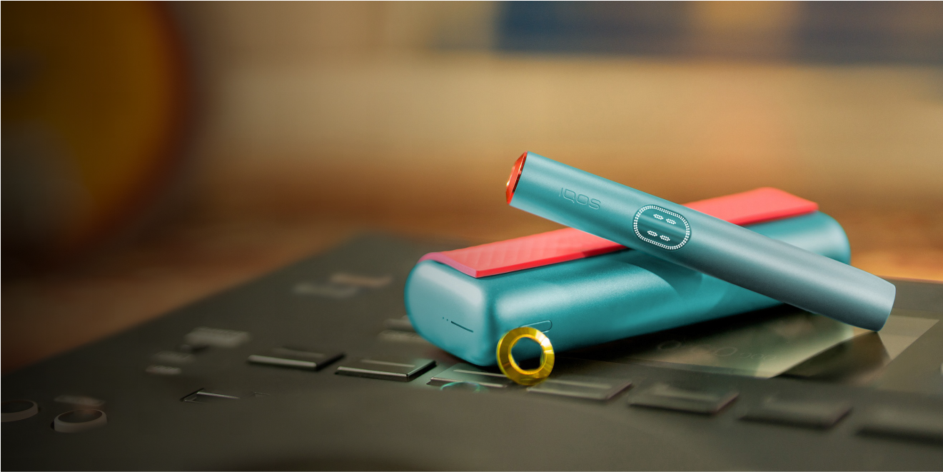 جهاز IQOS ILUMA i Prime باللون الأزرق المعدني مع لمسات حمراء، مع الحامل والشاحن على خلفية ضبابية.