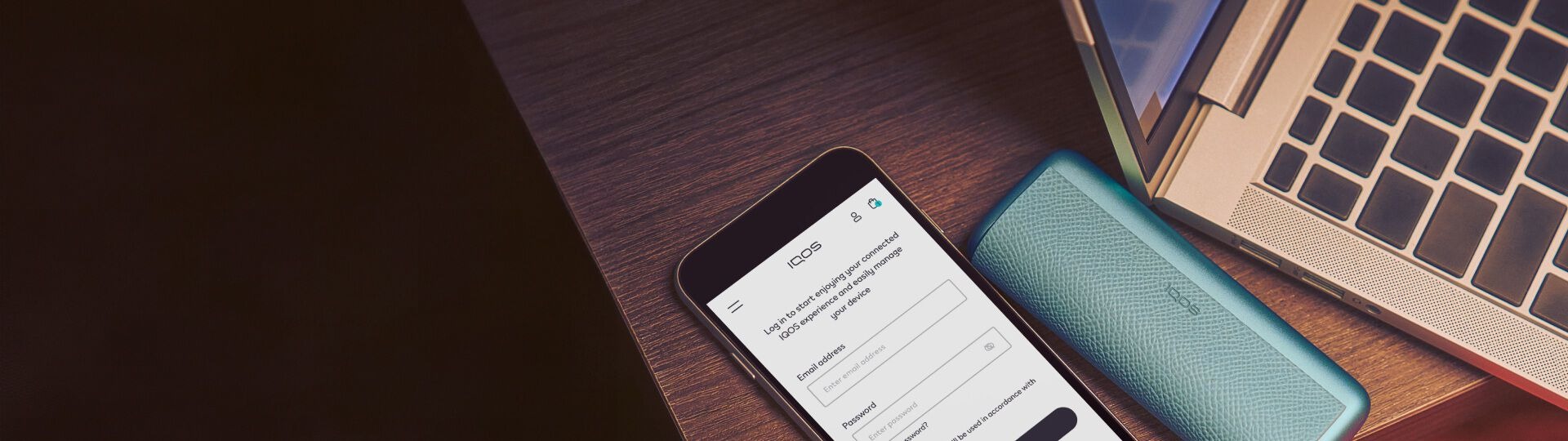 جهاز IQOS ILUMA i Prime باللون الأزرق الفيروزي موضوع بجانب هاتف ذكي يعرض شاشة تسجيل الدخول لتطبيق IQOS وحاسوب محمول على مكتب خشبي.