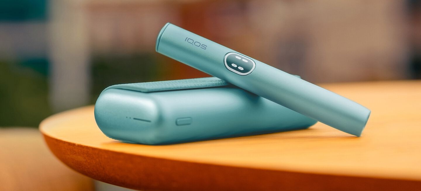 جهاز IQOS ILUMA i Prime باللون الفيروزي مع الحامل والشاحن موضوع على طاولة خشبية بخلفية ضبابية.
