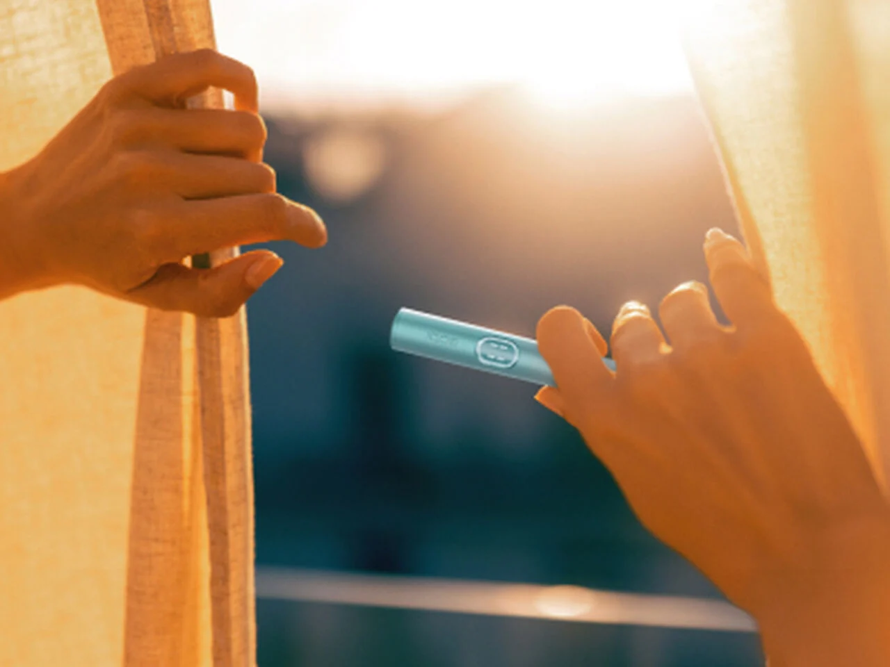يدان تمسكان جهاز IQOS ILUMA i Prime باللون الأزرق المعدني قرب ستارة مع أشعة الشمس في الخلفية.
