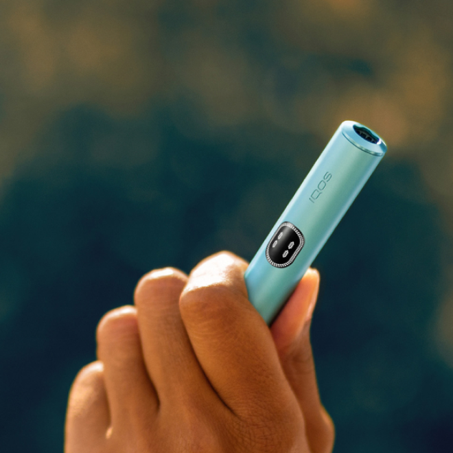 Hand holding a Breeze Blue IQOS ILUMA i device in a leafy background