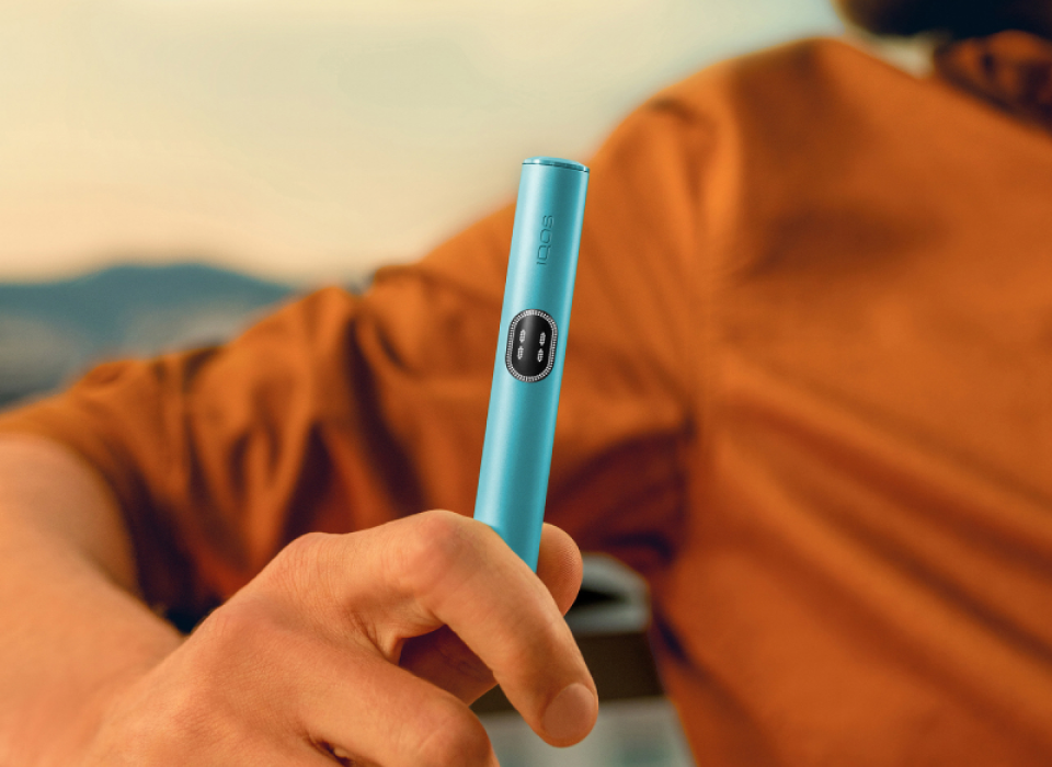 Hand holding a breeze Blue IQOS ILUMA i device, on a balcony.