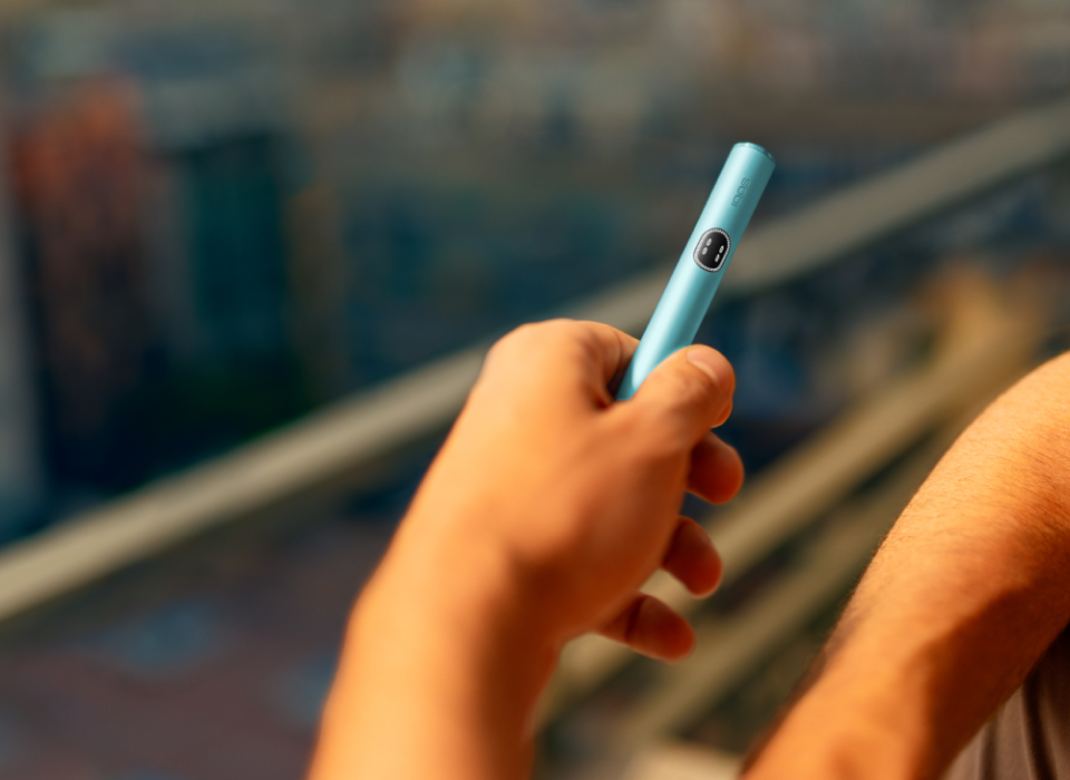 Hand holding a breeze Blue IQOS ILUMA i device, with the city as a background.