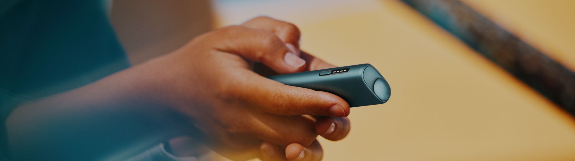 Mano sosteniendo un IQOS Iluma i ONE azul con fondo cálido desenfocado.