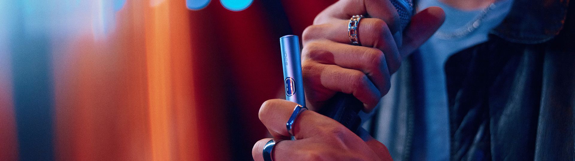 Mano usando un IQOS Iluma i ONE azul con luces difusas en el fondo.