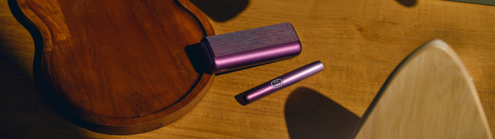 Dispositivo IQOS ILUMA i en color ciruela sobre una mesa de madera junto a un mate.