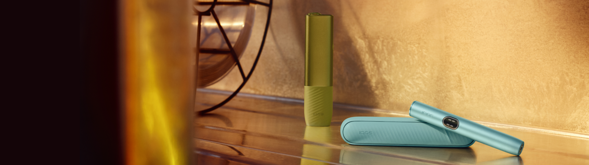 IQOS Iluma i Prime verde y un IQOS Iluma i azul sobre mesa iluminada con luz cálida.
