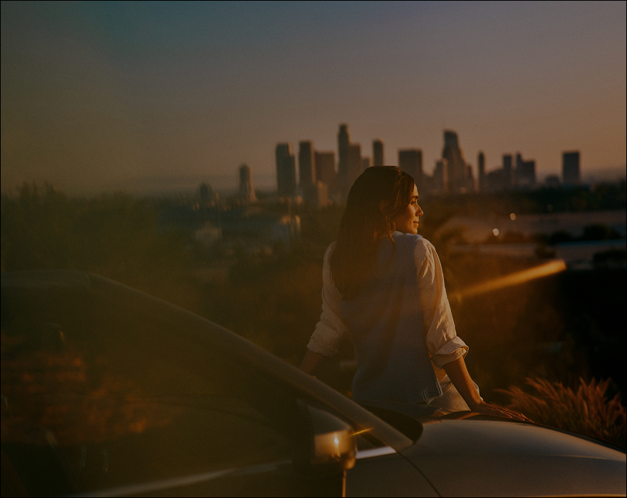 persona sentada sobre el cofre de un automóvil, mirando hacia un horizonte urbano al atardecer, con edificios iluminados por luz cálida