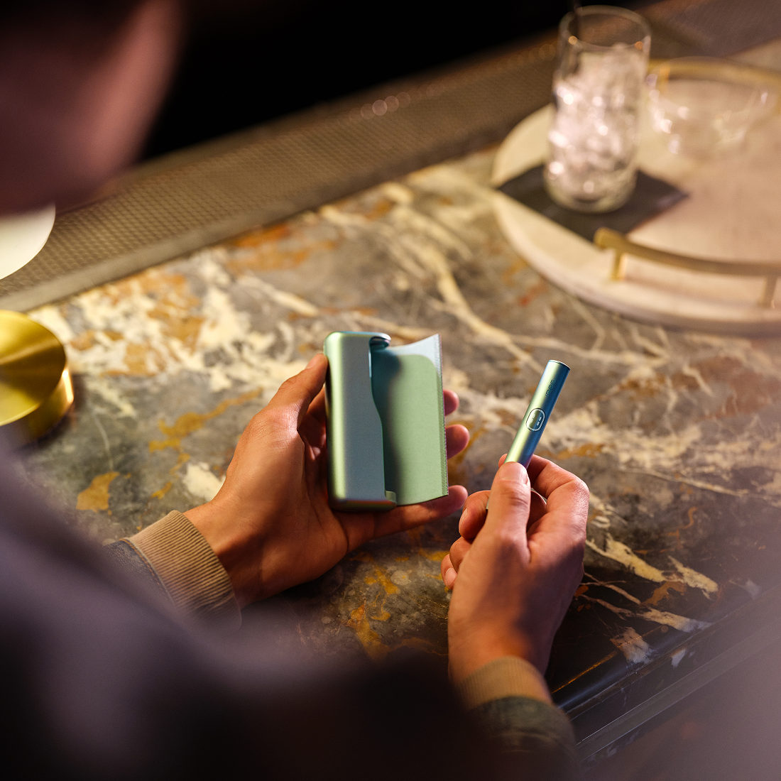 Persona colocando holder en dispositivo IQOS ILUMA PRIME color verde sobre mesa con accesorios.