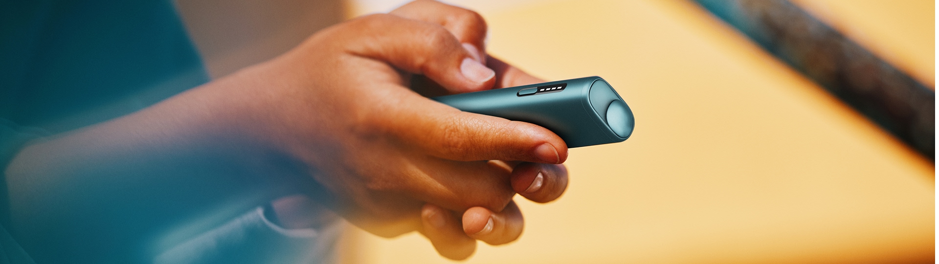 Persona usando IQOS Iluma i turquesa en exterior con fondo cálido desenfocado